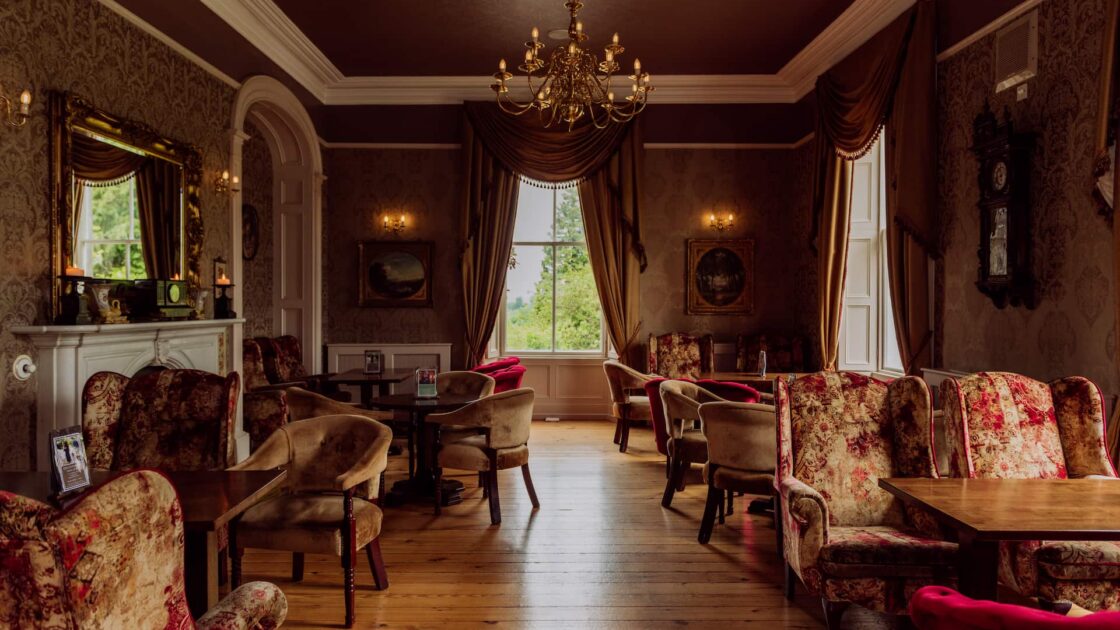 Elegant hotel lounge with vintage armchairs, ornate chandelier, and large windows offering a lush garden view.