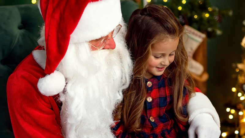 Santa Claus with a little girl in festive attire seated by a Christmas tree in a cosy hotel lobby, exuding holiday cheer.