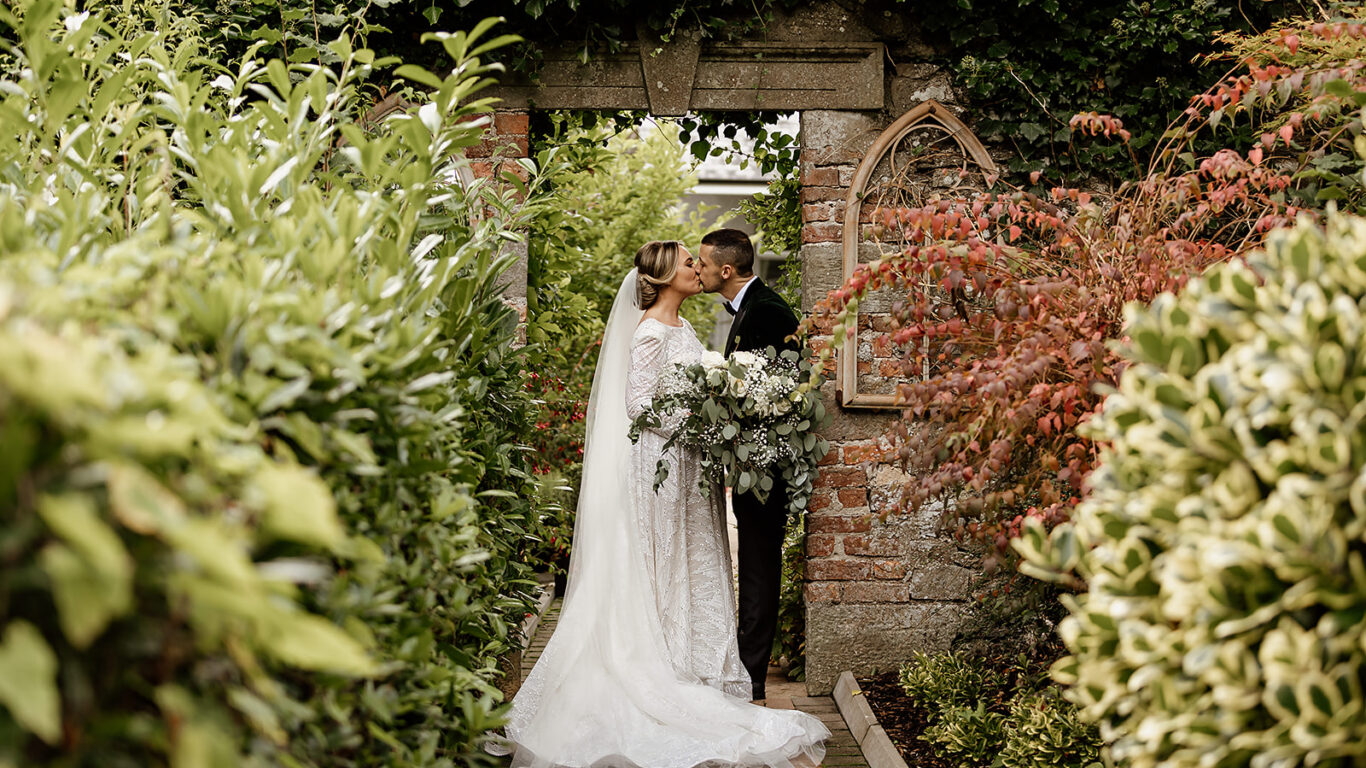 Romantic couple kiss in lush garden, green foliage, stone archway at luxury hotel, perfect for weddings and events.