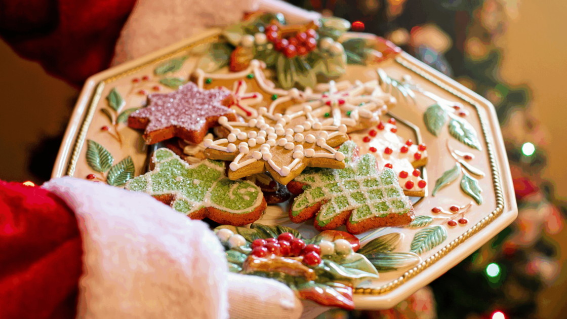 Festive gingerbread cookies on a decorative plate held by Santa, with a glowing Christmas tree in the background.