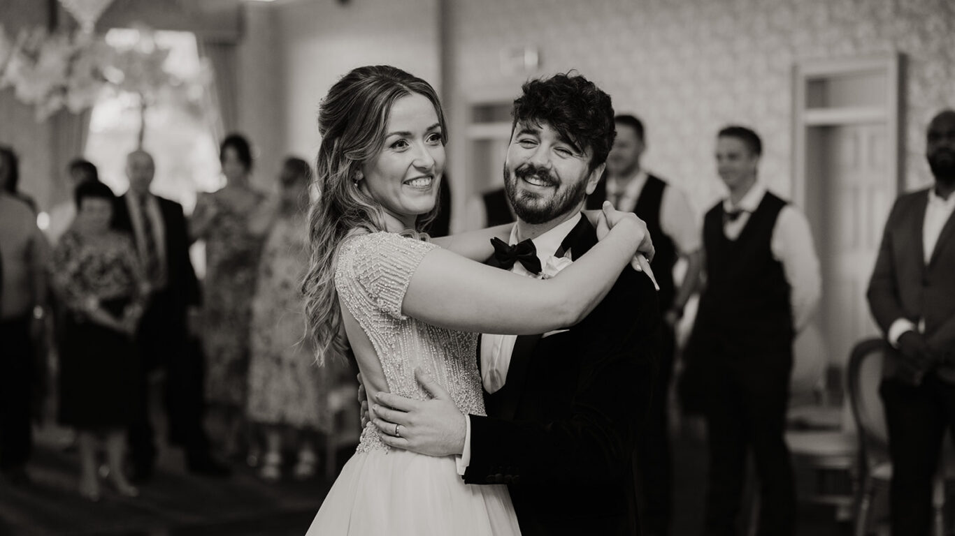 Newlyweds share a joyful dance in elegant ballroom with guests smiling and clapping, capturing a perfect wedding moment.