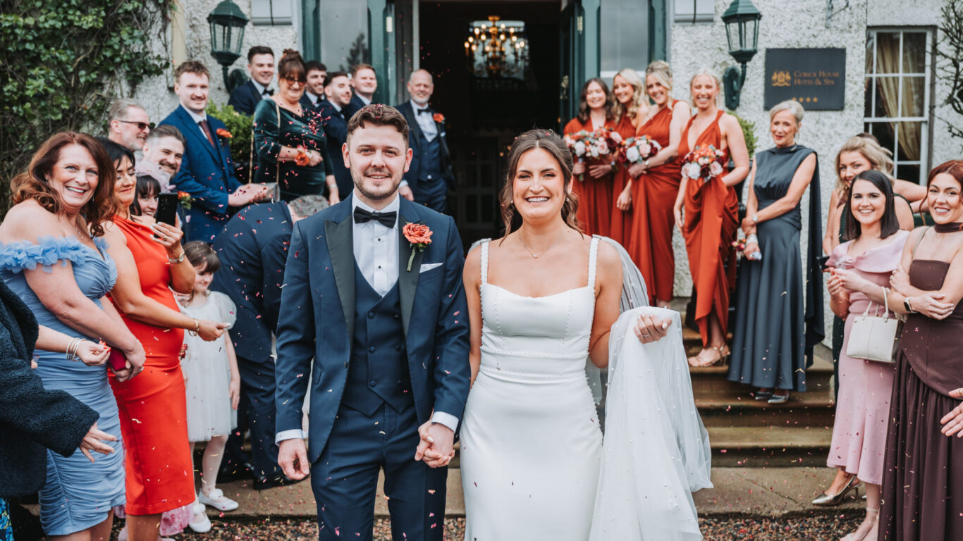 Happy couple celebrates wedding with guests at elegant countryside hotel entrance, surrounded by lush greenery and smiling faces.
