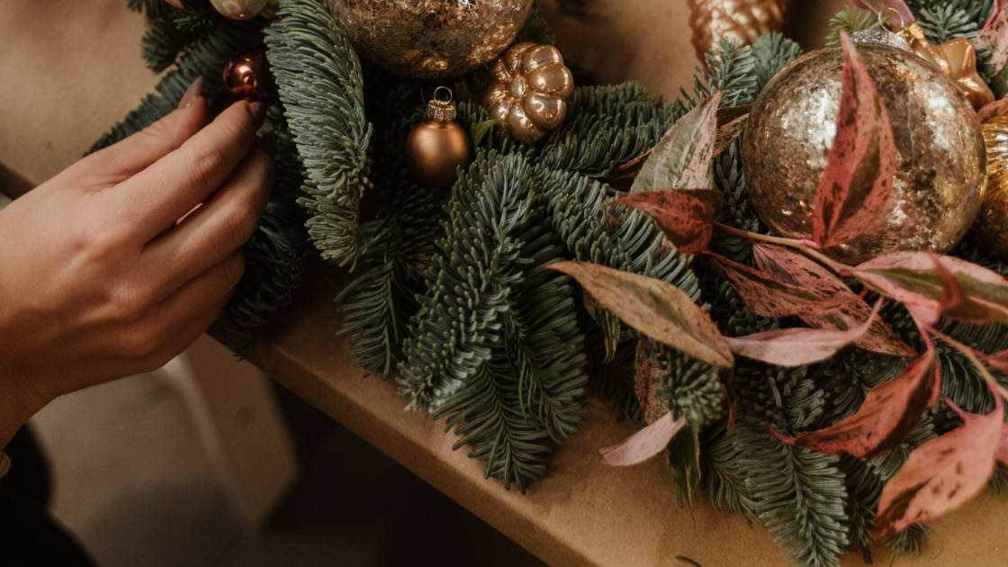 Hand arranging festive Christmas wreath with golden baubles at cosy hotel setting, evoking warm holiday ambiance.