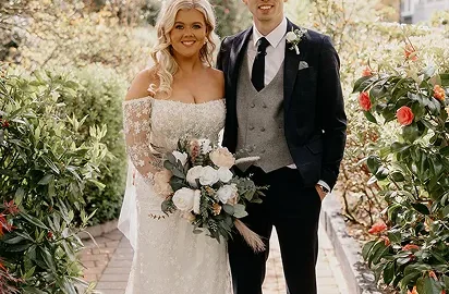 Couple in wedding attire stands on a brick path surrounded by lush, vibrant garden greenery, exuding romance and elegance.