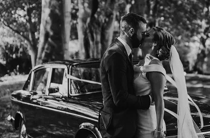 Romantic couple kissing by classic car under lush trees, perfect for dreamy weddings at our hotel venue.
