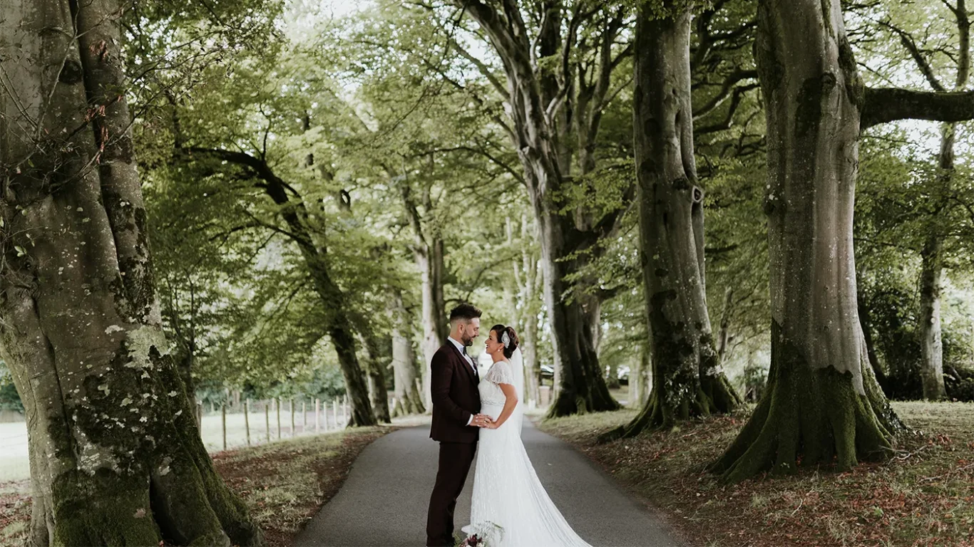 Charming couple in elegant wedding attire embrace on a tree-lined path at scenic hotel grounds, ideal for romantic getaways.