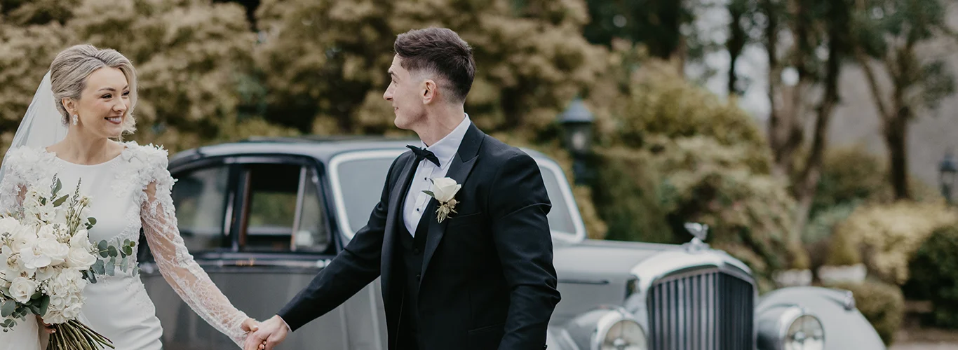 Newlyweds holding hands beside a classic car in lush hotel garden, perfect for romantic weddings and luxurious stays.